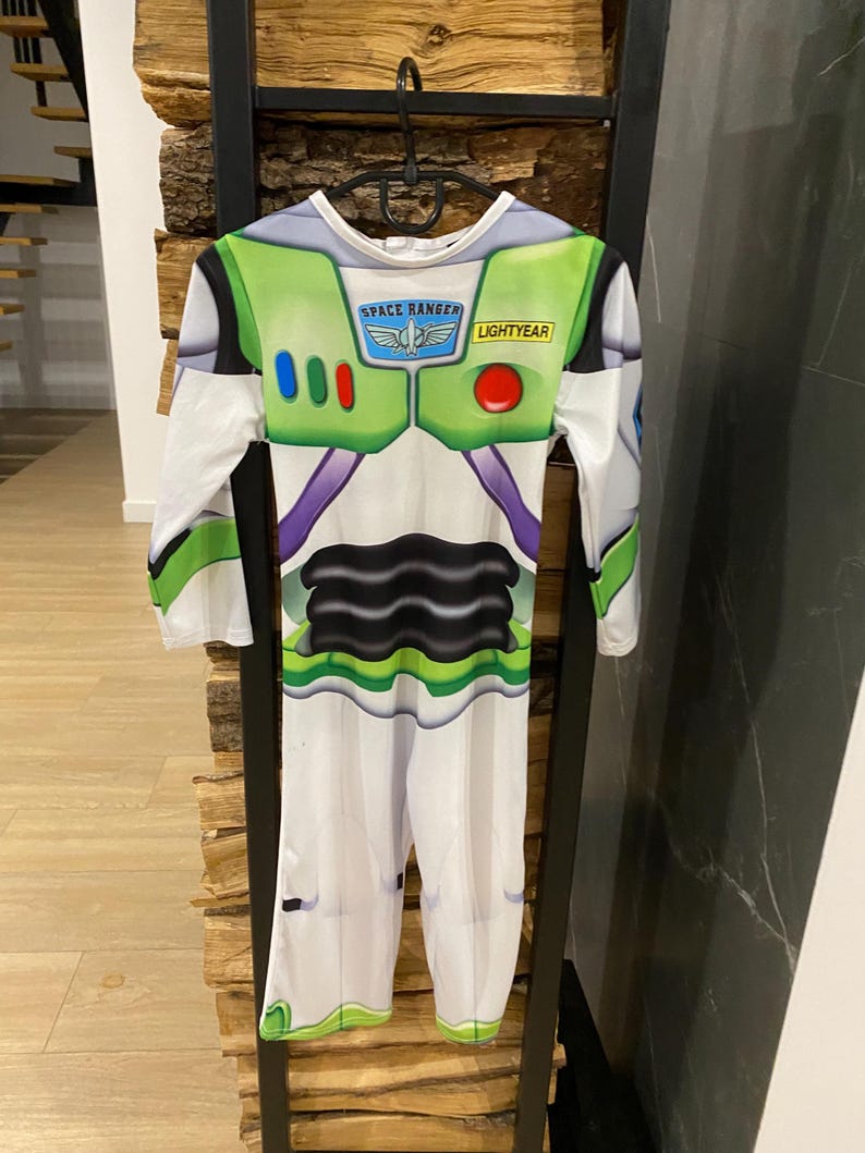 May include: A Buzz Lightyear costume with long sleeves and pants. The costume is primarily white with green, purple, and black accents. The chest area features the text "SPACE RANGER" and "LIGHTYEAR". The costume is hanging on a black hanger.