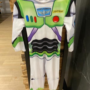 May include: A Buzz Lightyear costume with a white base, green accents, and the words "SPACE RANGER" and "LIGHTYEAR." The costume features a black and purple design on the torso and is hanging on a black hanger.