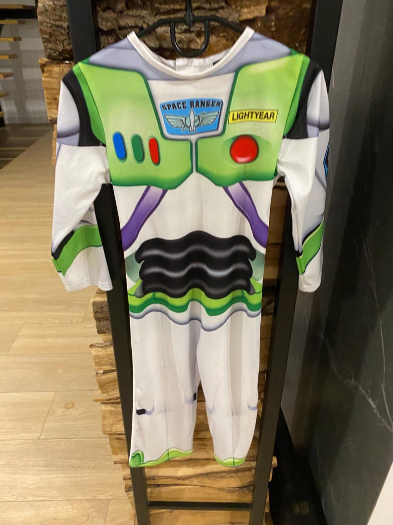 May include: A Buzz Lightyear costume with a white base, green accents, and the words "SPACE RANGER" and "LIGHTYEAR." The costume features a black and purple design on the torso and is hanging on a black hanger.