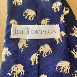 Op de afbeelding: Marineblauwe zijden stropdas met een herhalend patroon van beige olifanten. Het label leest "Jim Thompson". De olifanten zijn gestileerd en gelijkmatig verdeeld. Een klassiek accessoire. De stropdas is ongeveer 147 cm lang.