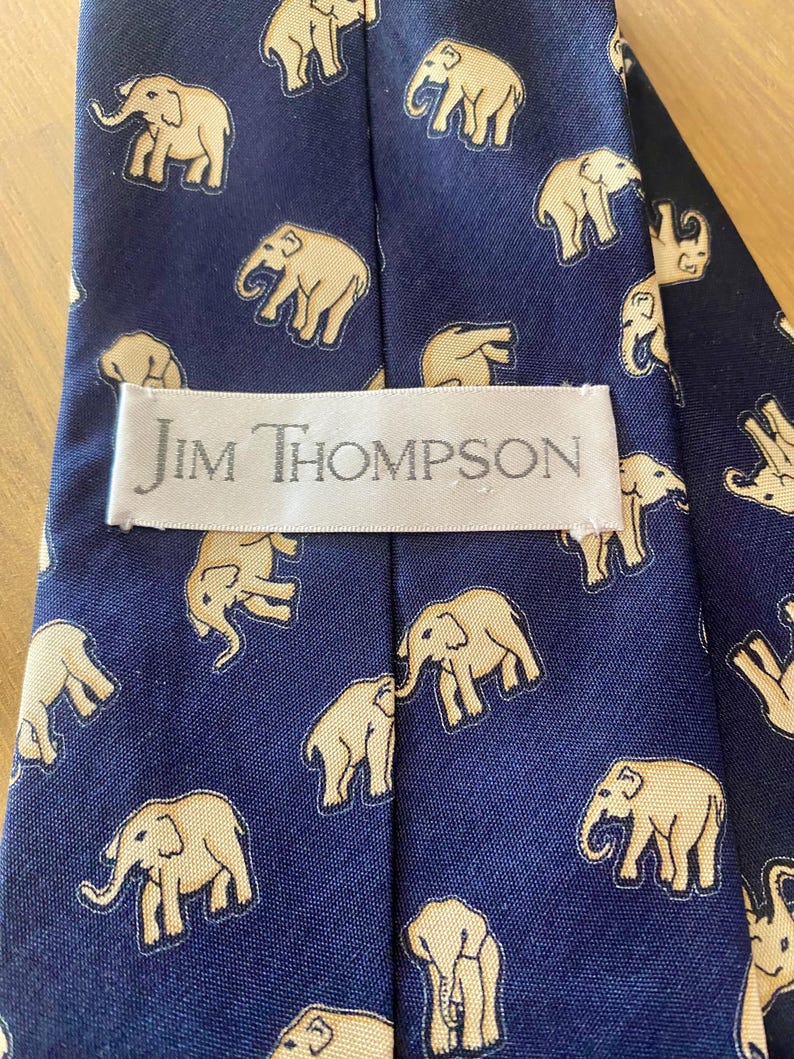 Op de afbeelding: Marineblauwe zijden stropdas met een herhalend patroon van beige olifanten. Het label leest "Jim Thompson". De olifanten zijn gestileerd en gelijkmatig verdeeld. Een klassiek accessoire. De stropdas is ongeveer 147 cm lang.