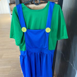 May include: A green t-shirt with blue overalls attached, featuring yellow buttons. The costume is hanging on a black hanger. The overall dress has a gathered skirt.