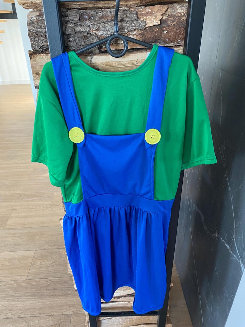 May include: A green t-shirt with blue overalls attached, featuring yellow buttons. The costume is hanging on a black hanger. The overall dress has a gathered skirt.