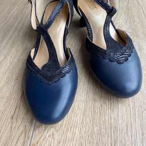 May include: A pair of navy blue T-strap shoes with a rounded toe and a scalloped edge detail. The shoes feature a textured black overlay on the T-strap and collar, a buckle closure, and a low heel. The interior insole is labeled with the brand name Hotter.