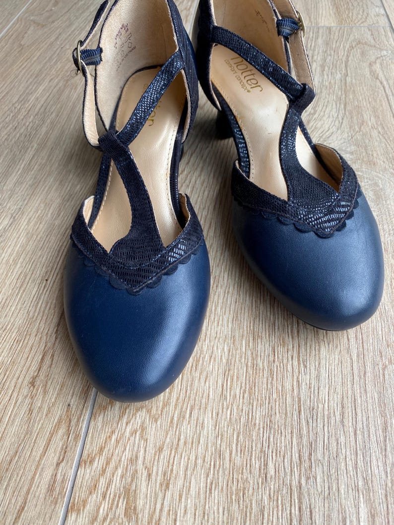 May include: A pair of navy blue T-strap shoes with a rounded toe and a scalloped edge detail. The shoes feature a textured black overlay on the T-strap and collar, a buckle closure, and a low heel. The interior insole is labeled with the brand name Hotter.