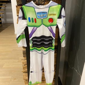May include: A Buzz Lightyear costume, featuring a white jumpsuit with green and purple accents. The chest area has a green panel with the words "SPACE RANGER" and "LIGHTYEAR". The costume has long sleeves and is hanging on a black hanger.