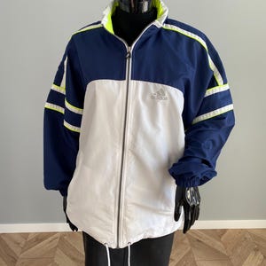Op de afbeelding: Een marineblauwe, witte en neongele Adidas-trainingsjas. De jas heeft een volledige ritssluiting, een trekkoord in de taille en het Adidas-logo op de borst. Witte strepen sieren de mouwen.