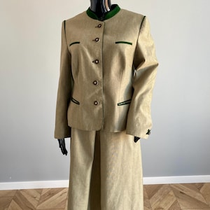 May include: A tan skirt suit with a long-sleeved jacket and a knee-length skirt. The jacket has a round collar and green trim. The suit is displayed on a black mannequin.
