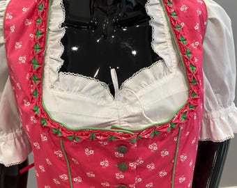 May include: A traditional pink dirndl dress with white floral patterns and green trim. The dress features a white blouse with ruffled sleeves and a square neckline. The bodice has decorative buttons.