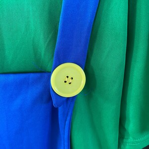 May include: Close-up of a green and blue costume. A bright yellow button with two holes is attached to a blue strap. The strap is sewn onto a green fabric. The costume appears to be made of a lightweight material.