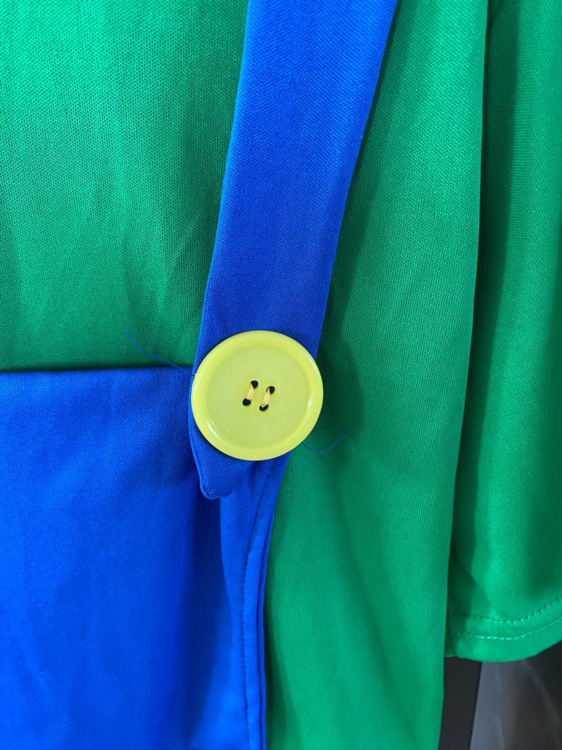 May include: Close-up of a green and blue costume. A bright yellow button with two holes is attached to a blue strap. The strap is sewn onto a green fabric. The costume appears to be made of a lightweight material.