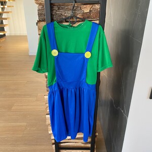 May include: A green t-shirt with blue overalls, featuring yellow buttons. The outfit is hanging on a black metal rack. The overall dress has a pleated skirt.