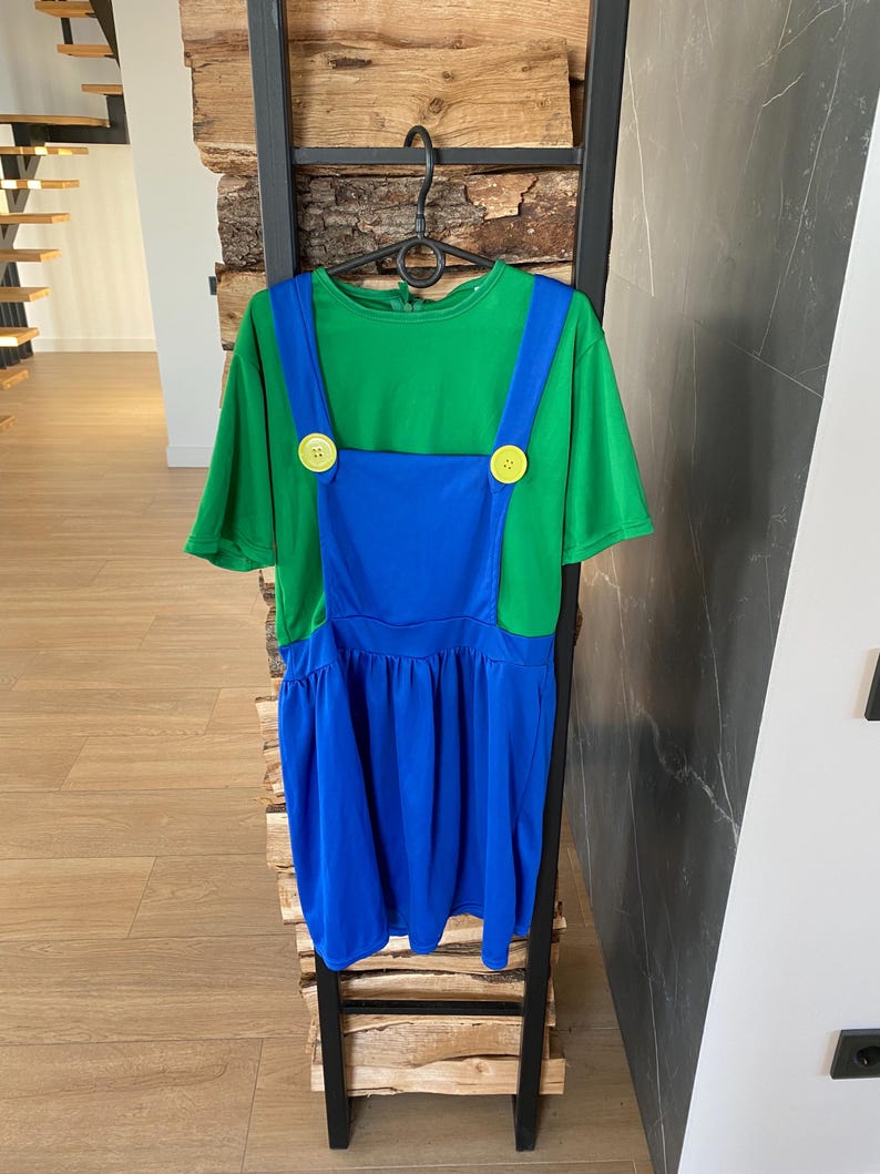 May include: A green t-shirt with blue overalls, featuring yellow buttons. The outfit is hanging on a black metal rack. The overall dress has a pleated skirt.