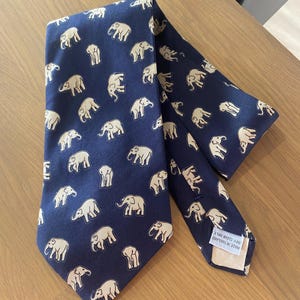 Op de afbeelding: Marineblauwe stropdas met een herhaald patroon van beige olifanten. De stropdas is gemaakt van een gladde stof en ligt op een houten oppervlak. De stropdas heeft een klassieke vorm, geschikt voor formele of zakelijke kleding.