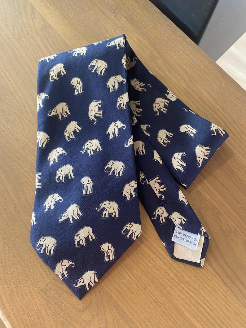 Op de afbeelding: Marineblauwe stropdas met een herhaald patroon van beige olifanten. De stropdas is gemaakt van een gladde stof en ligt op een houten oppervlak. De stropdas heeft een klassieke vorm, geschikt voor formele of zakelijke kleding.