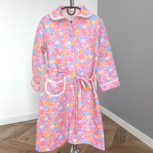 May include: A vintage floral print robe in pink, orange, blue, and yellow. The robe has a white lace trim on the collar, cuffs, and pocket. It is buttoned down the front and has a matching belt. The robe is displayed on a chrome hanger.