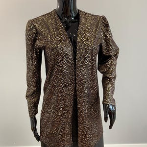 May include: A long-sleeved jacket featuring a black base with a dense pattern of small, metallic gold dots. The jacket has a V-neckline, structured shoulders with slight puff detailing, and a straight, open-front design that falls to the hips.