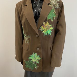 Könnte beinhalten: Ein brauner Blazer mit Blattapplikationen in Grün- und Regenbogenfarben. Der Blazer hat einen Zwei-Knopf-Verschluss und ein fallendes Revers. Die Blätter sind auf den Revers und der Vorderseite des Blazers angebracht.