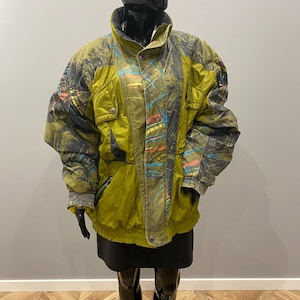 May include: A vintage olive green and gray ski jacket with a high collar and button closure. The jacket features colorful abstract patterns and two front pockets. The jacket is displayed on a black mannequin.