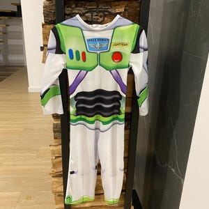 May include: A Buzz Lightyear costume with a white base and green, purple, and black accents. The costume features the Space Ranger logo and the word "Lightyear". The long-sleeved jumpsuit is hanging on a black metal rack.