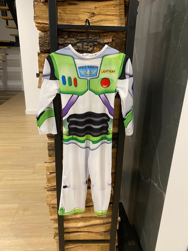 May include: A Buzz Lightyear costume with a white base and green, purple, and black accents. The costume features the Space Ranger logo and the word "Lightyear". The long-sleeved jumpsuit is hanging on a black metal rack.
