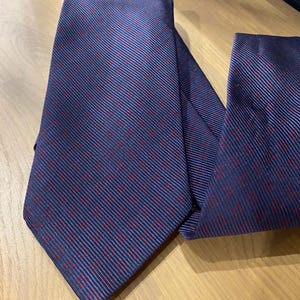 May include: A navy blue tie with a repeating pattern of thin red lines and dots. The tie is folded and lies on a light brown wooden surface. The tie's fabric has a subtle sheen.