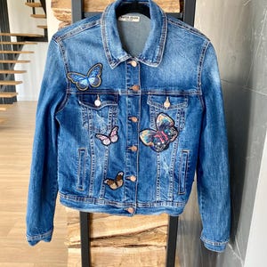 以下が含まれることがあります： 青いデニムジャケット。様々な色の蝶のパッチが付いています。 ジャケットはボタン留め、胸ポケットが2つあり、ラベルには「WATER JEANS」と書かれています。 黒い金属製のラックに掛けられています。