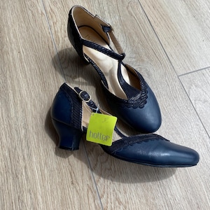 May include: A pair of navy blue T-strap heels with a low block heel and a scalloped edge detail. The shoes feature a textured fabric panel on the upper and a gold-tone buckle closure. A bright green tag with the word hotter is attached to one shoe.