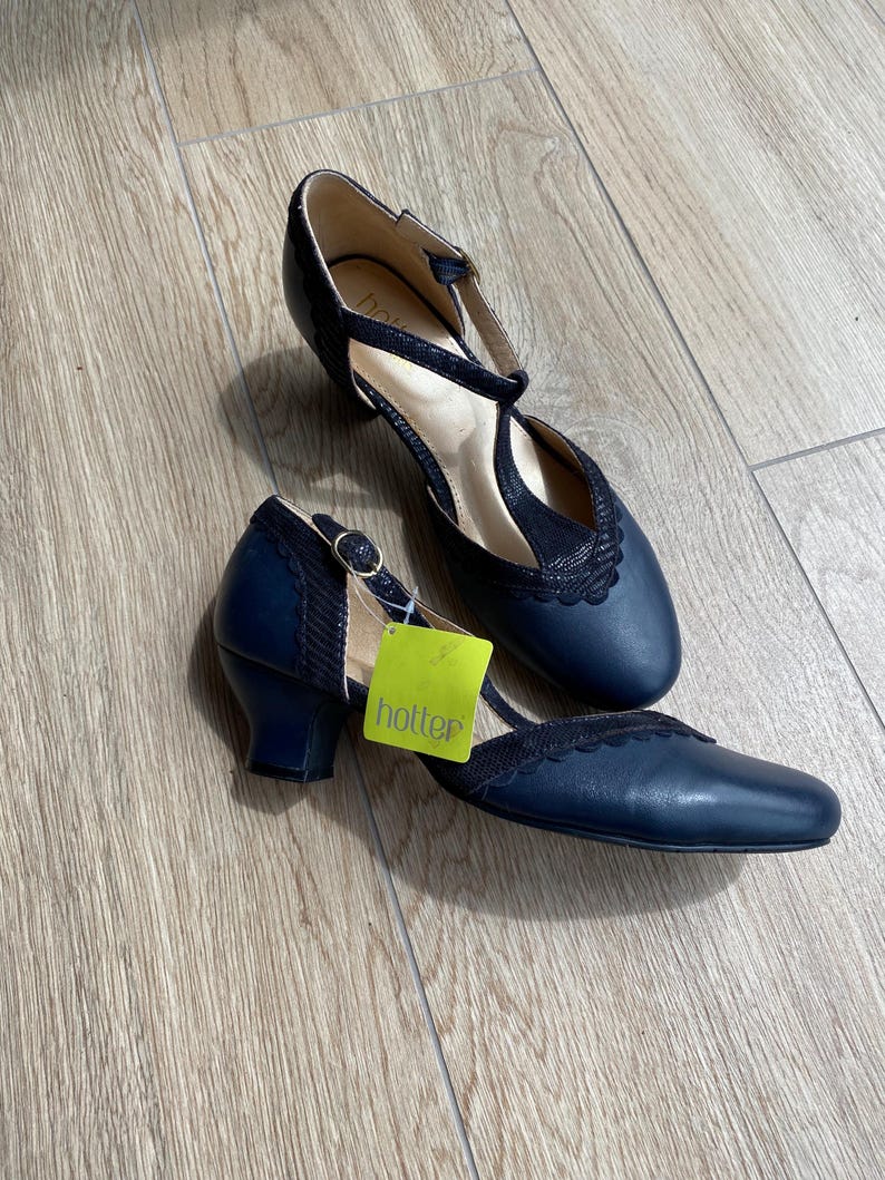 May include: A pair of navy blue T-strap heels with a low block heel and a scalloped edge detail. The shoes feature a textured fabric panel on the upper and a gold-tone buckle closure. A bright green tag with the word hotter is attached to one shoe.