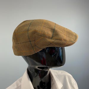 Puede incluir: Gorra plana de tweed marrón con un diseño a cuadros en amarillo y azul. La gorra se muestra sobre una cabeza de maniquí negra, combinada con una camisa blanca con cuello. Un sombrero clásico y elegante.