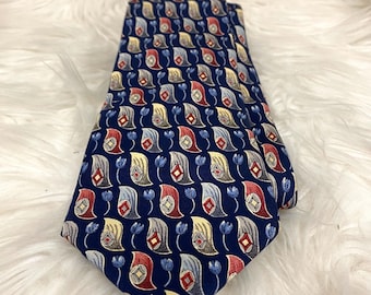 May include: Navy blue necktie with a repeating paisley pattern in red, yellow, and gray. The tie features small blue floral accents. The tie is folded and displayed on a white, fluffy surface.