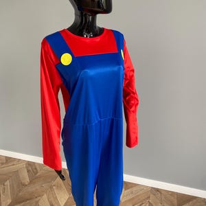 May include: A long-sleeved jumpsuit featuring a vibrant red shirt base with attached blue overalls. The blue section includes two prominent yellow circular buttons on the chest straps. The garment is displayed on a glossy black mannequin torso.