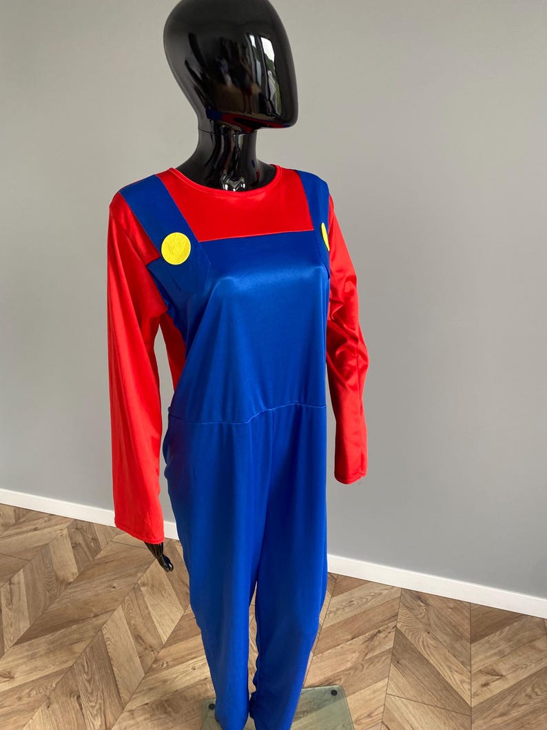 May include: A long-sleeved jumpsuit featuring a vibrant red shirt base with attached blue overalls. The blue section includes two prominent yellow circular buttons on the chest straps. The garment is displayed on a glossy black mannequin torso.
