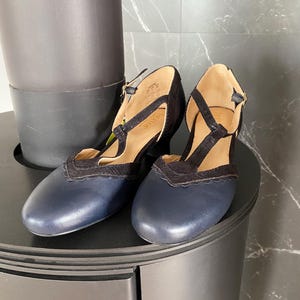 May include: A pair of navy blue leather T-strap shoes with black textured fabric overlays, scalloped edges, and a small block heel. The interior of the shoes features a tan lining with the brand name Hotter printed on the insole and a gold buckle.
