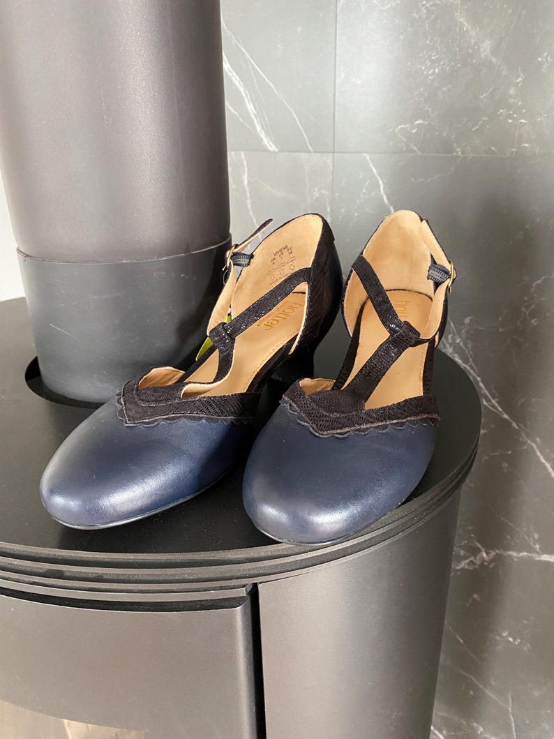 May include: A pair of navy blue leather T-strap shoes with black textured fabric overlays, scalloped edges, and a small block heel. The interior of the shoes features a tan lining with the brand name Hotter printed on the insole and a gold buckle.