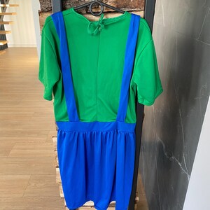 May include: A green and blue dress with short sleeves and suspender straps. The top is green, and the suspenders are blue. The skirt is a vibrant blue color. The dress is hanging on a black hanger.