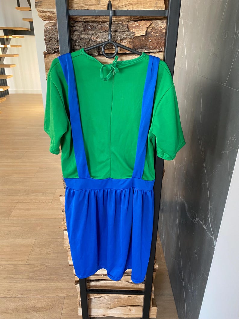 May include: A green and blue dress with short sleeves and suspender straps. The top is green, and the suspenders are blue. The skirt is a vibrant blue color. The dress is hanging on a black hanger.