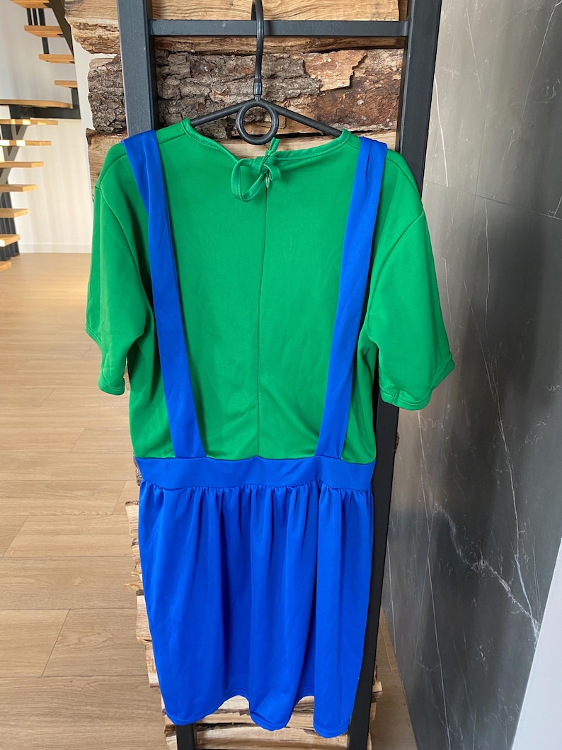 May include: A green and blue costume dress hanging on a black hanger. The top is green with blue suspenders, and the skirt is blue. The dress is made of a soft, lightweight fabric. The dress is designed to be worn as a costume.