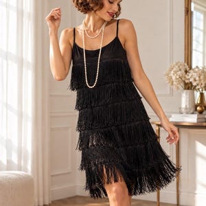 May include: A black sleeveless dress featuring multiple horizontal tiers of fringe, paired with a long white pearl necklace and a black sequined headband adorned with a black feather and a central circular decorative accent.