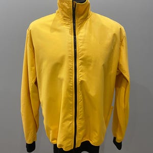 May include: A yellow track jacket with a high collar and a full-length black zipper. The jacket has black cuffs and a black bottom hem. The jacket is displayed on a mannequin.