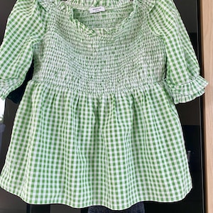 May include: Green and white gingham print top with a square neckline and ruffled trim. The top features a smocked bodice and three-quarter sleeves with ruffled cuffs. The fabric is a light, breathable material.