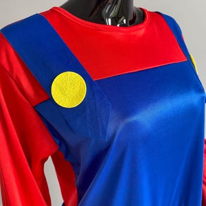 May include: A long-sleeved shirt featuring a red base with blue overall-style straps attached to the front. The straps are accented with large, circular yellow felt buttons on each side. The fabric has a smooth, slightly reflective finish.