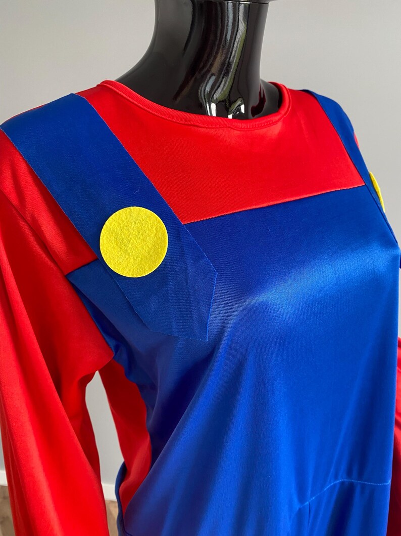 May include: A long-sleeved shirt featuring a red base with blue overall-style straps attached to the front. The straps are accented with large, circular yellow felt buttons on each side. The fabric has a smooth, slightly reflective finish.