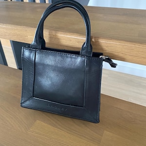 May include: A small, black leather handbag with a structured rectangular shape. The bag features a top handle and a zipper closure. The brand name "RADLEY" is subtly embossed on the front. The bag is sitting on a wooden surface.