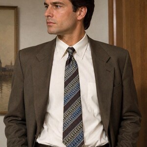 May include: A brown blazer worn over a crisp white collared shirt and a dark brown necktie featuring diagonal stripes in blue and yellow. The ensemble includes a dark brown leather belt with a rectangular buckle, completing the formal attire.