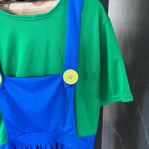 May include: A green t-shirt with a blue overall-style dress. The dress features blue straps, a blue bib, and a blue skirt. Large yellow buttons secure the straps to the green shirt. The outfit is hanging on a black hanger.