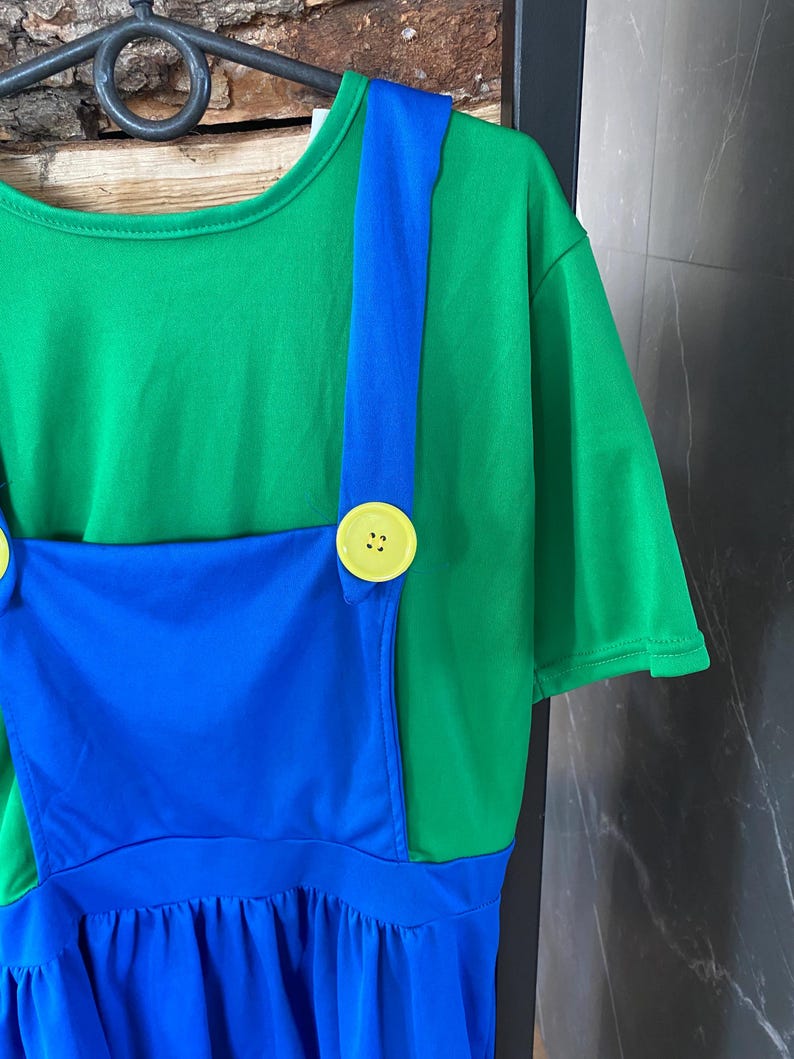 May include: A green t-shirt with a blue overall-style dress. The dress features blue straps, a blue bib, and a blue skirt. Large yellow buttons secure the straps to the green shirt. The outfit is hanging on a black hanger.