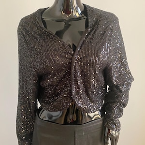 May include: A black sequined jacket with long sleeves and a V-neck design. The jacket is displayed on a black mannequin. The sequins give the jacket a shiny appearance, reflecting light. The jacket is a stylish piece of apparel.