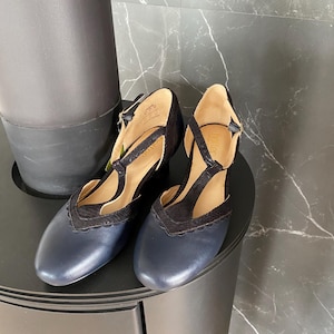 May include: A pair of navy blue and black T-strap shoes featuring a rounded toe, a scalloped edge detail along the vamp, and a buckled ankle strap. The shoes have a smooth leather-like finish on the toe and a textured fabric on the T-strap.