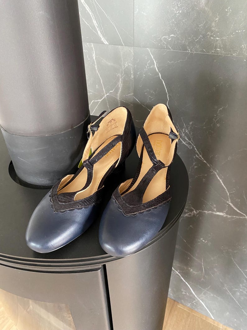 May include: A pair of navy blue and black T-strap shoes featuring a rounded toe, a scalloped edge detail along the vamp, and a buckled ankle strap. The shoes have a smooth leather-like finish on the toe and a textured fabric on the T-strap.